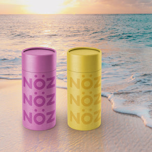 Two Noz reef safe sunscreen on the beach at sunset in the sand with waves lapping at the bottom edge of the sun cream bottles